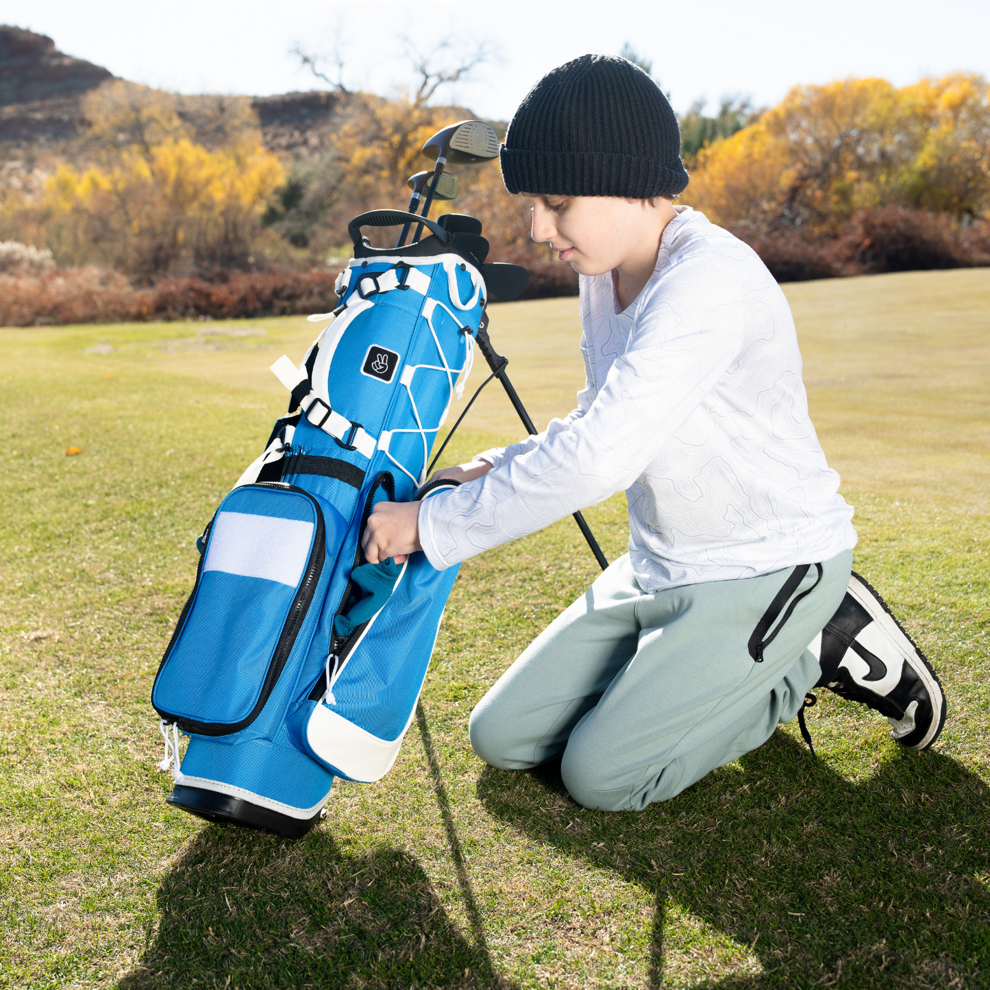 A young golfer wearing a black beanie and white long-sleeve shirt kneels on a golf course, adjusting the pocket of a blue & white Recess kids golf bag with a stand, featuring multiple compartments and golf clubs inside, set on a golf course