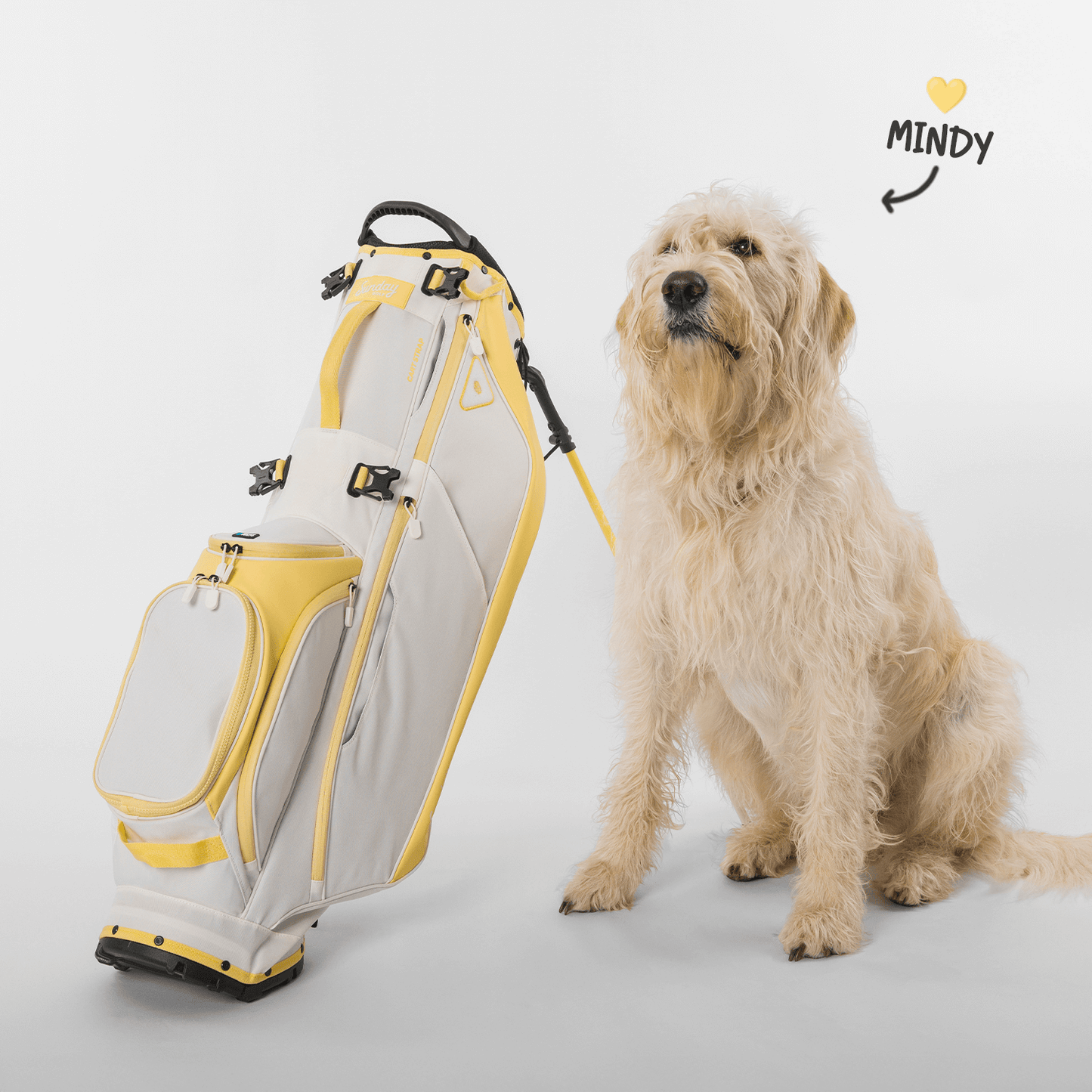 A full-size stand golf bag with white and lemon yellow colors, named 'Mindy', with a dog sitting next to it.