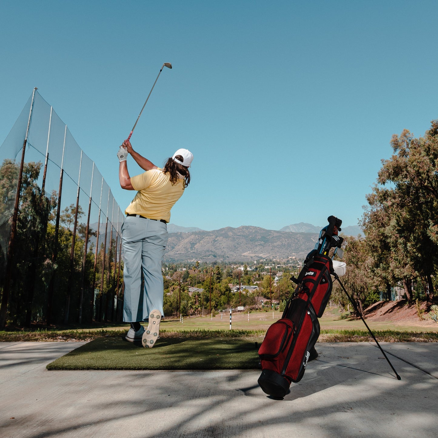 man swinging golf club at driving range with deep red burgundy loma xl golf bag by sunday golf