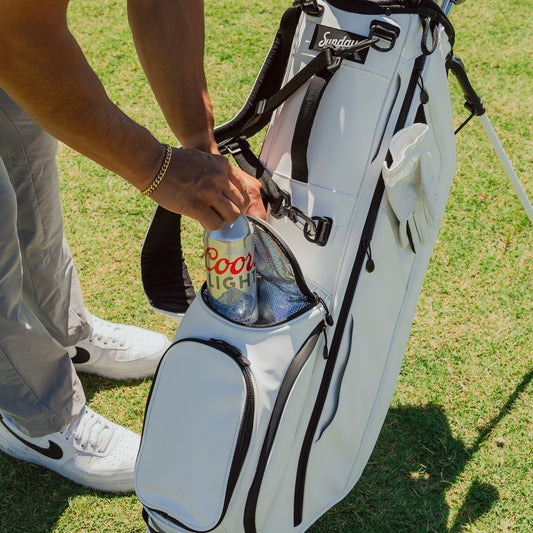Golfer placing a drink into insulated cooler pocket of a white golf stand bag on grass course